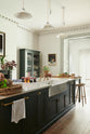 An oblique view of a long kitchen island with a double sink, an aged brass Ionian tap, and a curiosity cupboard in the grand bakehouse green classic English kitchen. Adorned with loaves of bread, leeks, red grapes, potted plants, still life paintings, and homemade jams; the room feels homey and welcoming despite its large and decorative appearance.