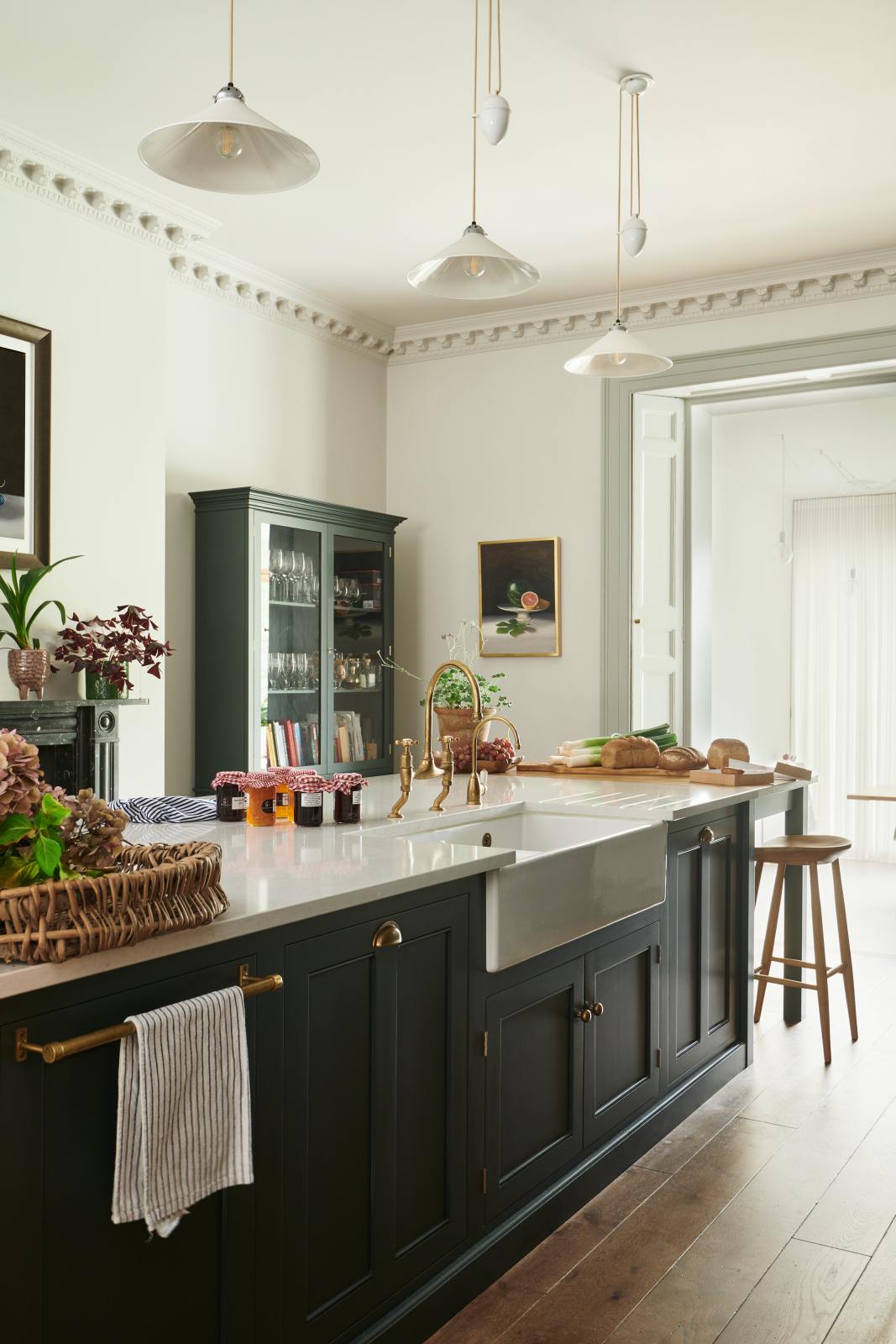 An oblique view of a long kitchen island with a double sink, an aged brass Ionian tap, and a curiosity cupboard in the grand bakehouse green classic English kitchen. Adorned with loaves of bread, leeks, red grapes, potted plants, still life paintings, and homemade jams; the room feels homey and welcoming despite its large and decorative appearance.