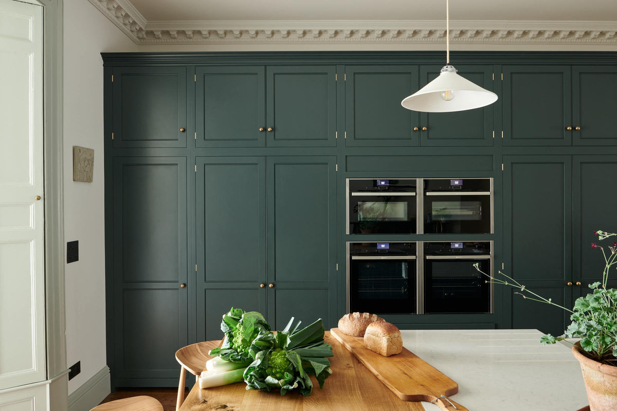 Looking at the edge of a large full-height pantry cupboard, oven cabinets, fridge/freezer cupboard, and a long kitchen island with a double sink, an aged brass Ionian tap, Silestone worktop and a solid oak worktop prep table in the grand bakehouse green classic English kitchen. Adorned with loaves of bread, leeks, romanesco cauliflower, and potted plants, the room feels homey and welcoming despite its large and decorative appearance.