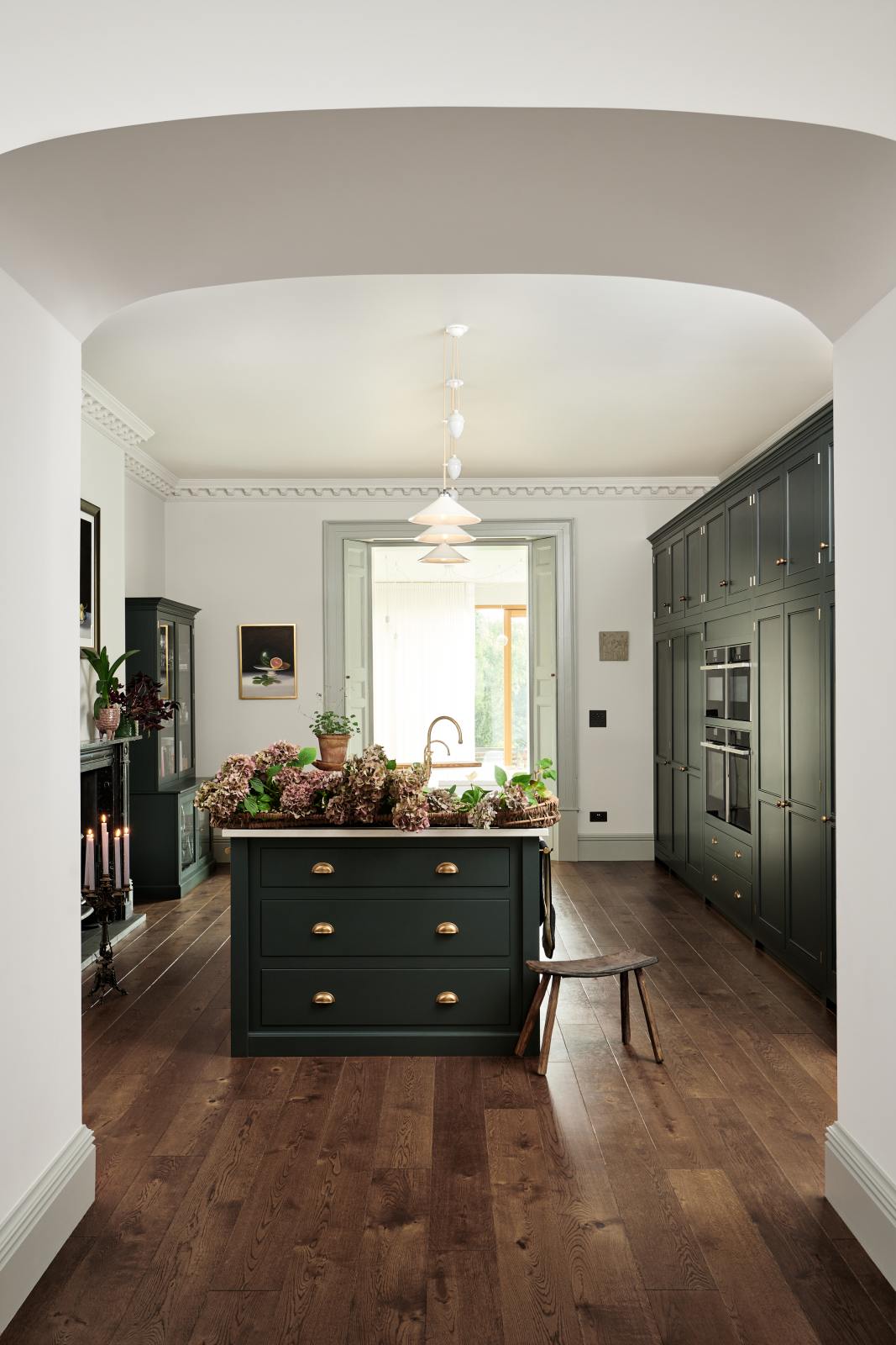 Looking through a large archway is a grand, Bakehouse green classic English kitchen has large full-height pantry cupboards, oven cabinets, fridge/freezer cupboards, a long kitchen island with a double sink, an aged brass Ionian tap, and a curiosity cupboard. Adorned with white and blush pink hydrangeas, romanesco cauliflower, potted plants, still life paintings, a candelabra, and homemade jams, the room feels homey and welcoming despite its large and decorative appearance.