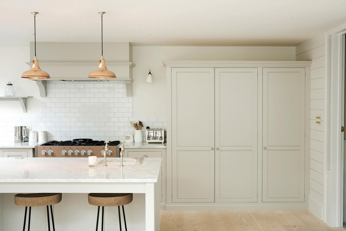 This simple and stylish classic English kitchen features a sink and cooker run, a suspended false chimney breast, a large pantry cupboard, and a kitchen island topped with Carrara marble. Beautifully decorated with light copper pendant lights, a silver toaster, a silver kettle, white ceramics, and white splashback tiles, it creates a functional and beautiful kitchen space.