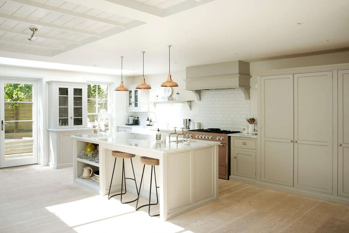 A simple and stylish classic English kitchen features a sink and cooker run, a suspended false chimney breast, a wall cupboard, a glazed larder, and a kitchen island topped with Carrara marble. Beautifully decorated with light copper pendant lights, a silver toaster, a silver kettle, muted ceramics, and white splashback tiles, it creates a functional and beautiful kitchen space.