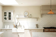 This close-up of a simple and stylish classic English kitchen highlights the sink and cooker run, featuring a double Belfast sink, an antique silver Ionian tap, and a Carrara marble worktop. It includes a glazed wall cupboard and a shelf. Beautifully decorated with a light copper pendant light, glass containers, white ceramics, a silver kettle, wooden cleaning equipment, and white splashback tiles, this space creates a functional and beautiful kitchen atmosphere.