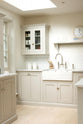 An oblique view of a simple and stylish classic English kitchen’s double Belfast sink with antique silver Ionian tap, Carrara marble worktop, glazed wall cupboard, and shelf. Beautifully decorated with light copper rimmed clock, glass containers, wooden cleaning equipment, and white splashback tiles, it creates a functional and beautiful kitchen space.
