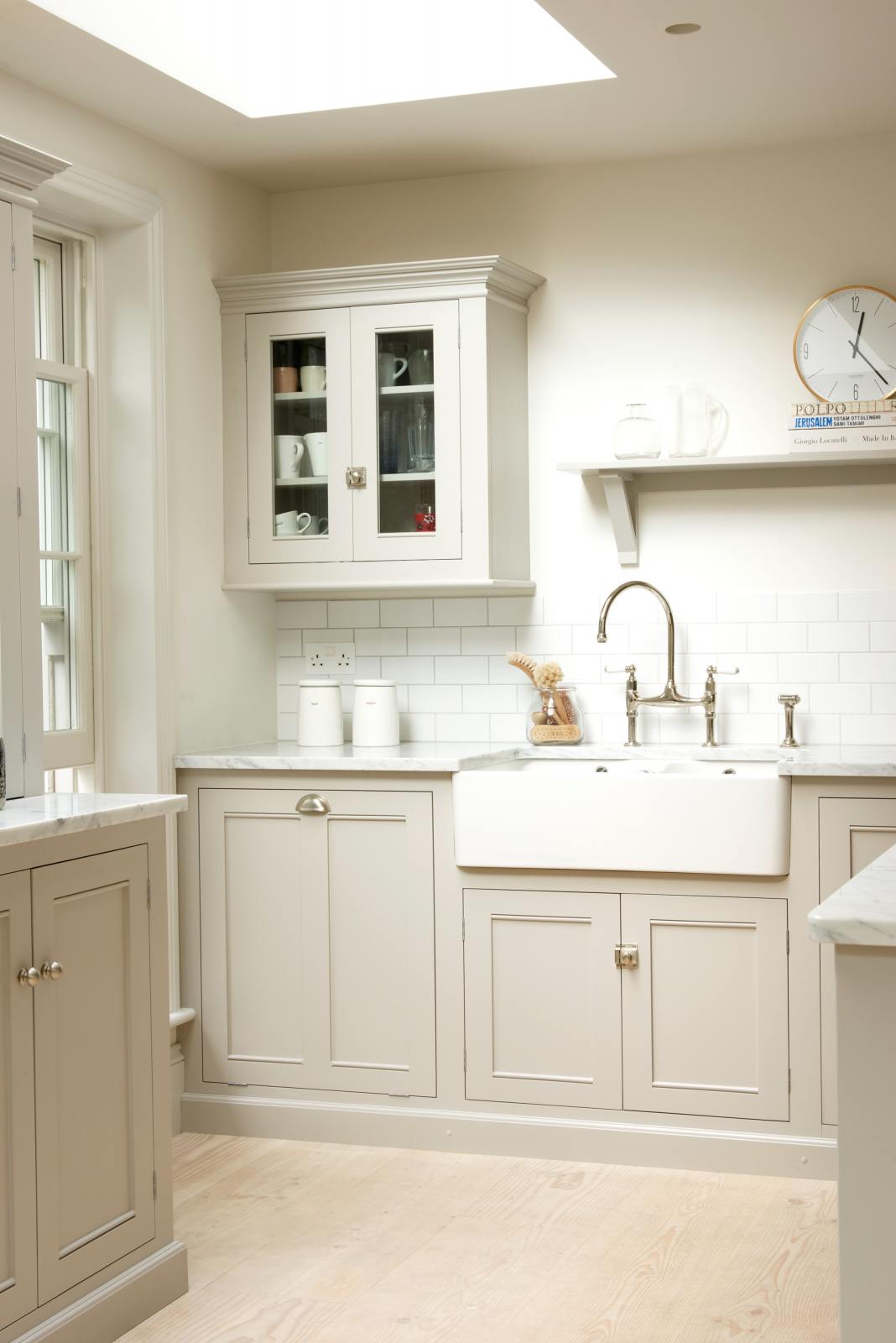 An oblique view of a simple and stylish classic English kitchen’s double Belfast sink with antique silver Ionian tap, Carrara marble worktop, glazed wall cupboard, and shelf. Beautifully decorated with light copper rimmed clock, glass containers, wooden cleaning equipment, and white splashback tiles, it creates a functional and beautiful kitchen space.