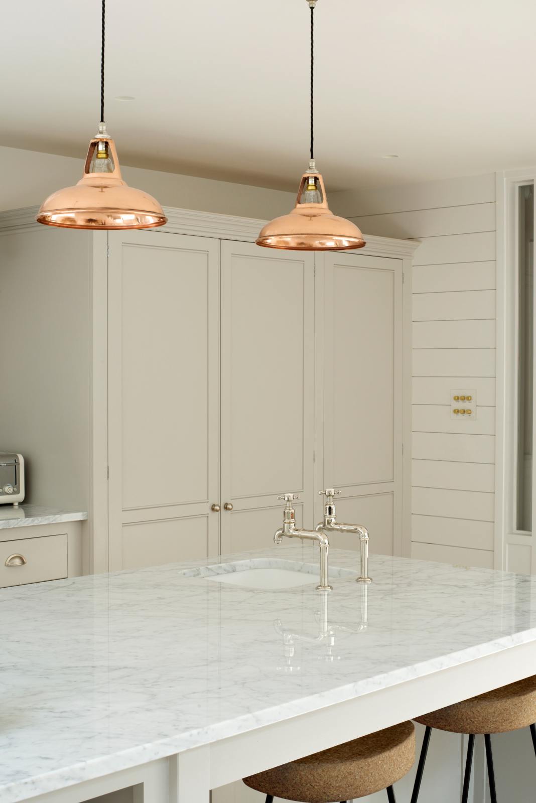 An oblique view of a simple and stylish classic English kitchen features a large pantry cupboard, and a kitchen island topped with Carrara marble. Beautifully decorated with light copper pendant lights, a silver toaster, antique silver Mayan taps, and cork stools, it creates a functional and beautiful kitchen space.