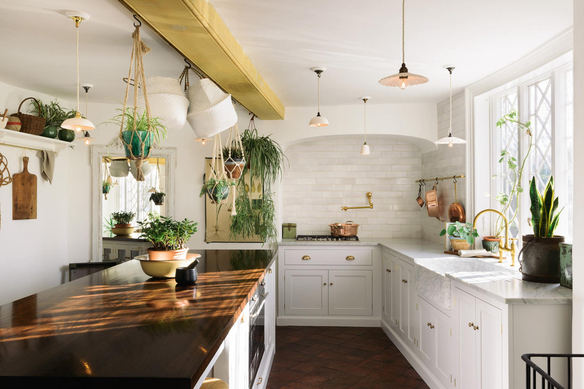 The white linen, classic English kitchen features an L-shaped sink and cooker run with Carrara marble worktop, a large single Carrara marble sink, an aged brass Ionian tap, pot filler tap and white crackle metro splashback tiles. A large kitchen island with a solid oak worktop adds character. Lush green hanging plants and white porcelain lights, with the original yellow beam, enhance the bright and cheerful atmosphere, making the kitchen lively and inviting.