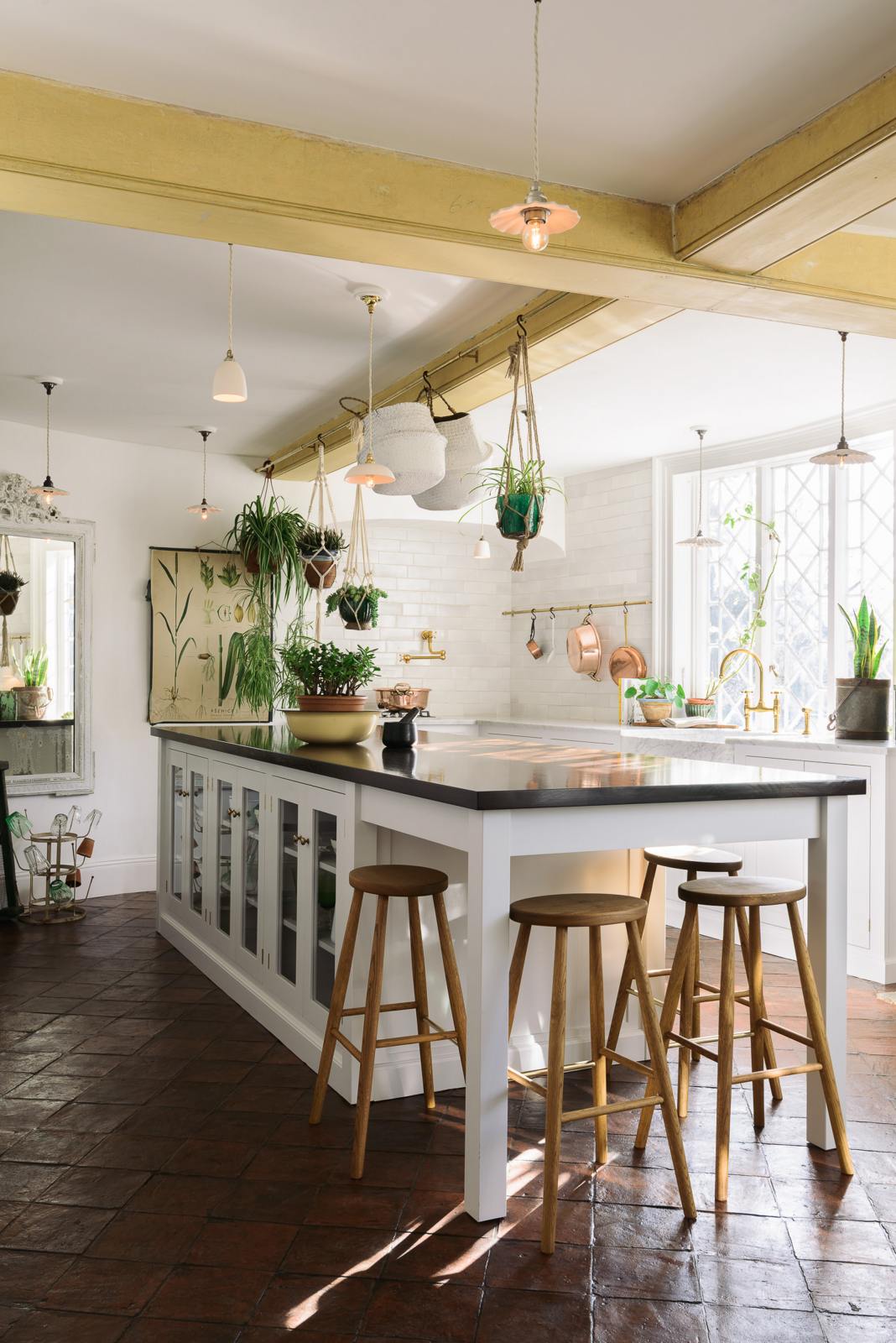 The white linen, classic English kitchen features an L-shaped sink and cooker run with Carrara marble worktop, a large single Carrara marble sink, an aged brass Ionian tap, and white crackle metro splashback tiles. In the centre is a large kitchen island with a solid oak worktop, breakfast nook and Helen stools. Lush green hanging plants and white porcelain lights, with the original yellow beam, enhance the bright and cheerful atmosphere, making the kitchen lively and inviting.