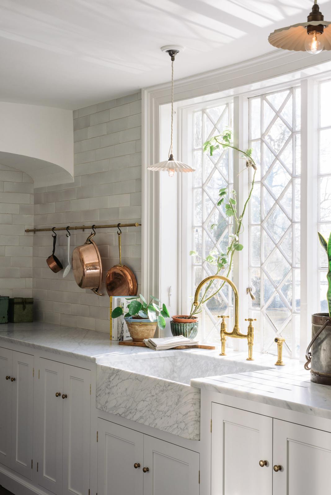 A close and angled look at a white linen, classic English kitchen features an L-shaped sink and cooker run with Carrara marble worktop, a large single Carrara marble sink, an aged brass Ionian tap, and white crackle metro splashback tiles. Lush green potted plants, white porcelain lights, and aged brass hanging rails with copper pots and pans enhance the bright and cheerful atmosphere, making the kitchen lively and inviting.