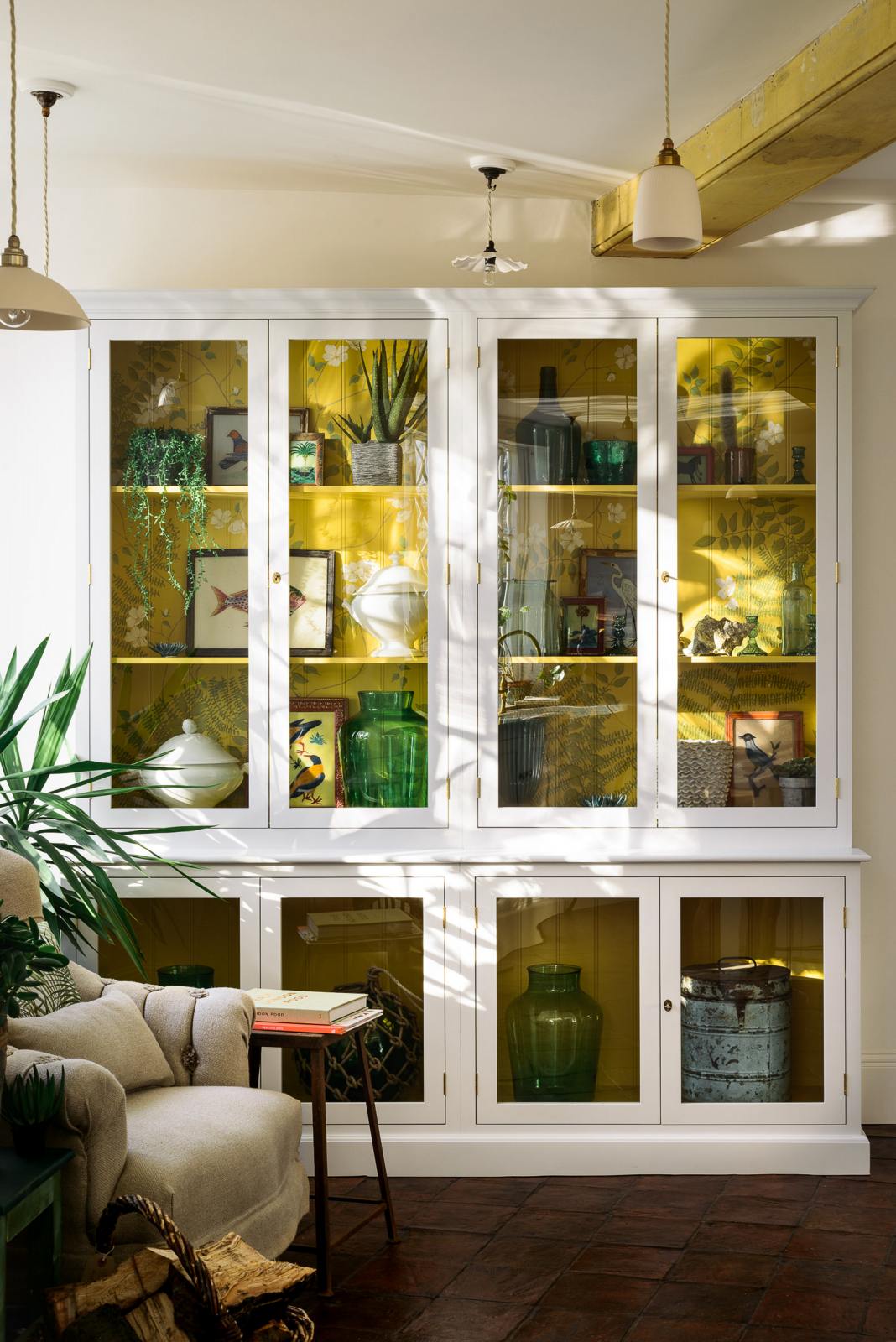 A large white linen curiosity cupboard with a yellow interior features hand-painted white flowers and green foliage, paying homage to the original yellow beams of the historical Millhouse building. It showcases beautiful vintage ceramics, glass centrepieces, framed detailed illustrations, and potted plants.