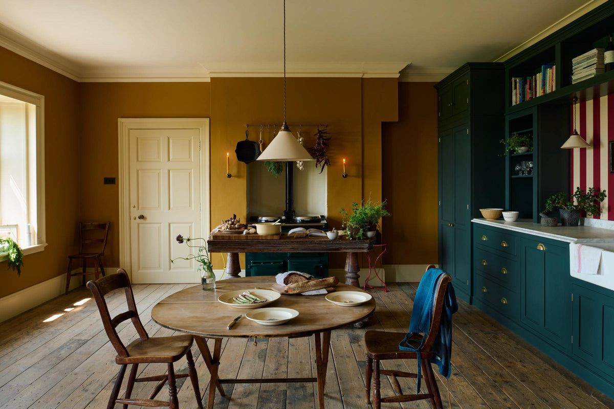 A lovely modern kitchen that retains the charm of its original structure and flooring, using the three primary colours in a muted, mature palette. The spacious kitchen features a Clerkenwell Blue full-height pantry cabinet, a long sink run with a marble worktop, and an open countertop cabinet and shelf. An emerald green stove sits within the chimney breast, accompanied by a deVOL cream pendant light above a vintage round wooden table, with matching chairs and a prep table.