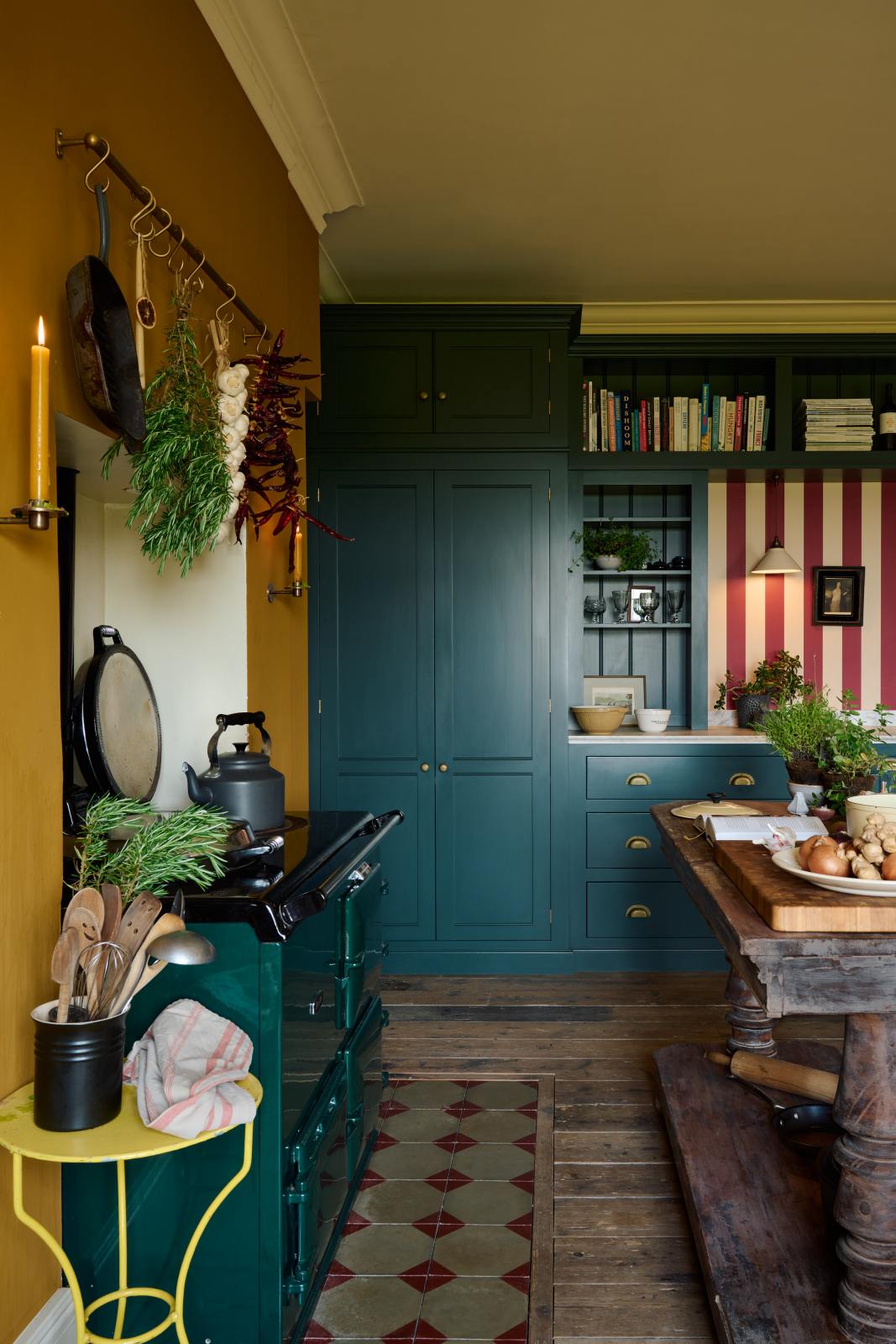 Nestled between the emerald green gas stove—set within the chimney breast with an aged brass hanging rail for herbs, garlic, and chillies—and the large vintage oak prep table, are the Clerkenwell Blue full-height pantry cabinet, a long sink run with a marble worktop, an open countertop cabinet, a high shelf, and a red-and-white striped splashback.