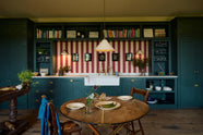 A lovely modern kitchen that retains the charm of its original structure and flooring, using the three primary colours in a muted, mature palette. The spacious kitchen features Clerkenwell Blue full-height pantry cabinets, a long sink run with a marble worktop, an aged brass Ionian tap, and an open countertop cabinet with a high shelf. It's completed by a deVOL cream pendant light hanging above a vintage round wooden table and matching chairs.