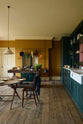 A lovely modern kitchen that retains the charm of its original structure and flooring, using the three primary colours in a muted, mature palette. The spacious kitchen features a Clerkenwell Blue full-height pantry cabinet, a long sink run with a marble worktop, and an open countertop cabinet and shelf. An emerald green stove sits within the chimney breast, accompanied by a deVOL cream pendant light above a vintage round wooden table, with matching chairs and a prep table.