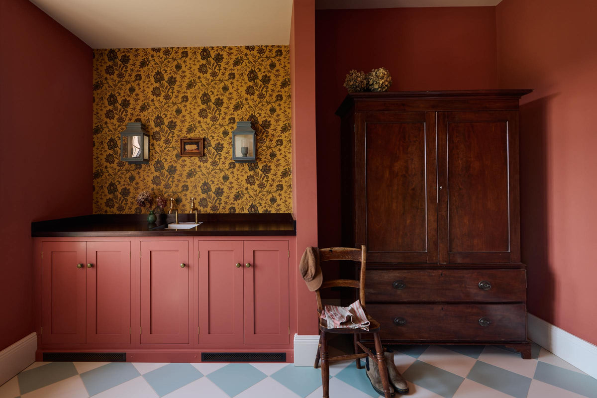 A small scullery room with a light red sink run, polished sapele worktop, a single sink fitted with aged brass Mayan taps, strikingly detailed floral wallpaper, and light blue and white porcelain floor tiles. Beside the concave sits a rich oak dresser with drawers, a chair, and a bunch of yellow flowers.