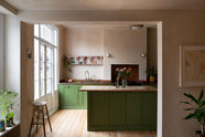 A romantic, decorative, Parisian-inspired green classic English kitchen featuring an L-shaped sink and cooker run with an aged copper worktop, an aged brass Ionian tap, and an oak worktop kitchen island. Above the sink, a pink vintage-framed shelf is adorned with small colourful abstract prints, lace runners, a bouquet of blush-brown chrysanthemums, and a plate of grapefruit and dragon fruit.