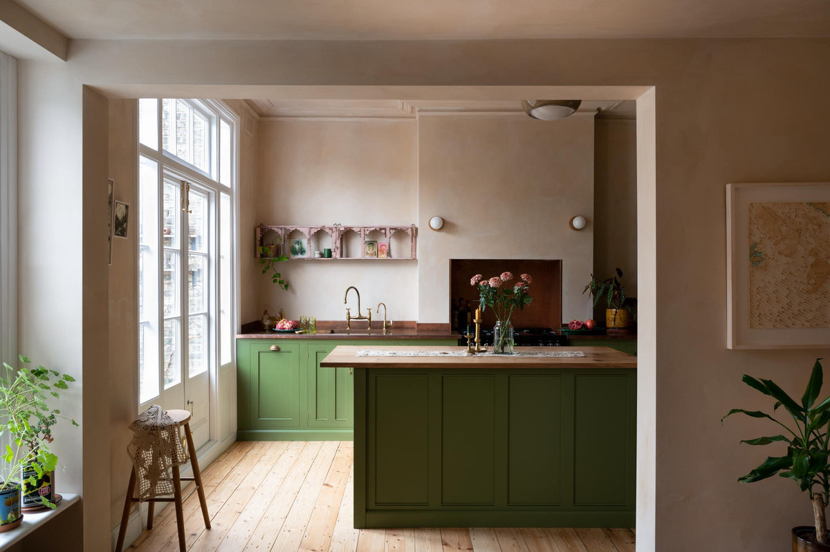 A romantic, decorative, Parisian-inspired green classic English kitchen featuring an L-shaped sink and cooker run with an aged copper worktop, an aged brass Ionian tap, and an oak worktop kitchen island. Above the sink, a pink vintage-framed shelf is adorned with small colourful abstract prints, lace runners, a bouquet of blush-brown chrysanthemums, and a plate of grapefruit and dragon fruit.