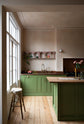 A romantic, decorative, Parisian-inspired bean green classic English kitchen featuring an L-shaped sink and cooker run with an aged copper worktop, an aged brass Ionian tap, and an oak worktop kitchen island. Above the sink, a pink vintage-framed shelf is adorned with small colourful abstract prints, lace runners, a bouquet of blush-brown chrysanthemums, and a strikingly red plate of dragon fruits.