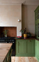 A closer look at a romantic, decorative, Parisian-inspired green classic English kitchen, featuring an L-shaped sink and cooker run with an aged copper worktop and an oak worktop kitchen island. Adorned with lush green house plants, and a strikingly red plate of grapefruits with brown, marble-washed walls and ceiling.