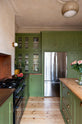 Nestled in between a romantic, decorative, Parisian-inspired green classic English kitchen’s L-shaped sink and cooker run with an aged copper worktop and an oak worktop kitchen island. There’s a large full-length cabinet with closed top cupboards, built-in stainless steel fridge/freezer and reeded glazed countertop cupboards. Adorned with lush green house plants, a bouquet of blush-brown chrysanthemums, and a strikingly red plate of grapefruits.