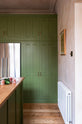 Nestled between a romantic, decorative, Parisian-inspired bean green classic English kitchen’s oak worktop kitchen island and entrance. There is a large full-length cabinet with closed upper cupboards and a built-in stainless steel fridge/freezer. It is adorned with a bouquet of blush-brown chrysanthemums, lace runners, and a wooden framed artwork.