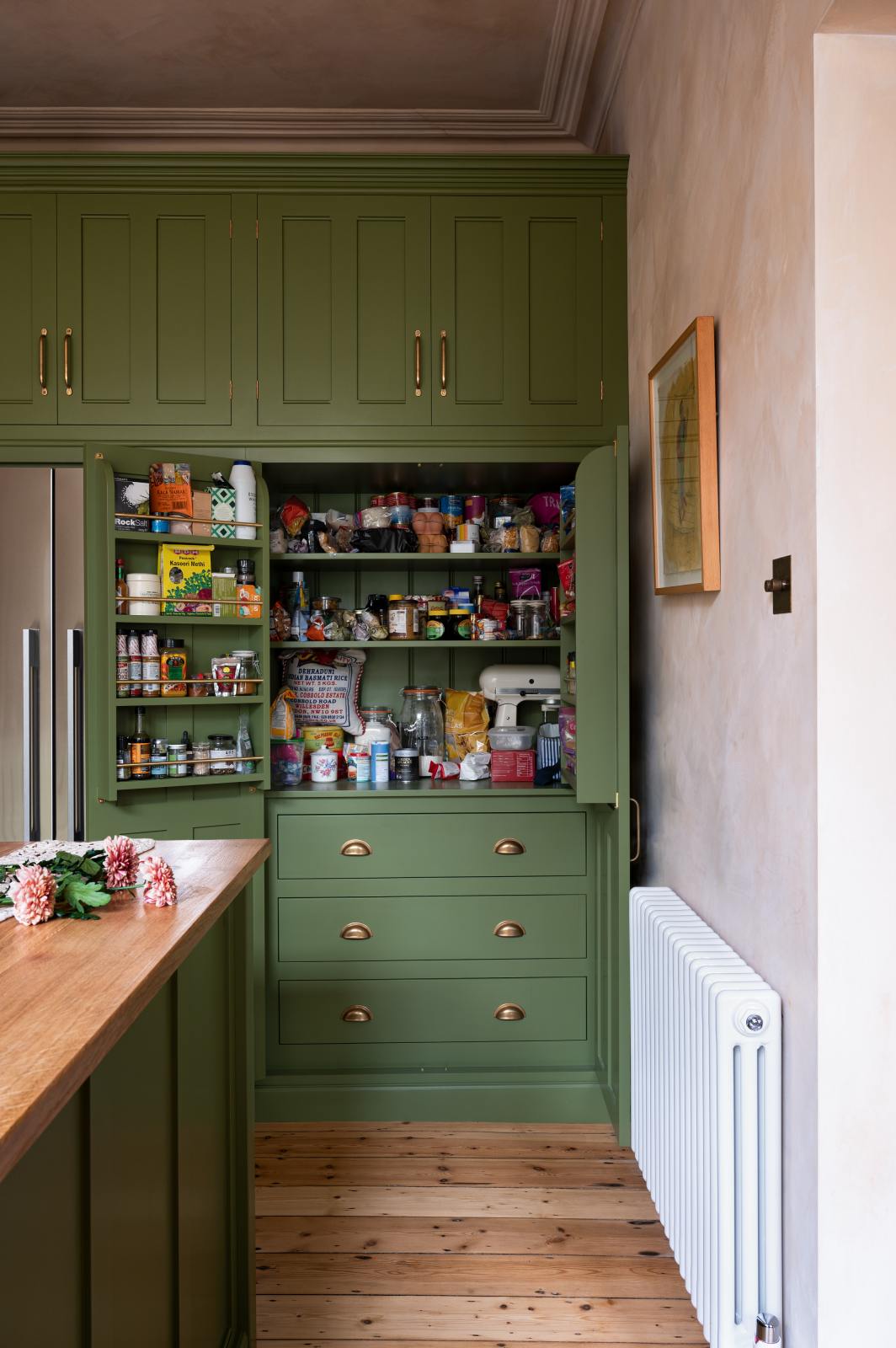 Nestled between the oak worktop kitchen island and the entrance of a romantic, decorative, Parisian-inspired classic English green kitchen is a large, open full-length cabinet with closed top cupboards and a built-in stainless steel fridge-freezer. The cabinet is fully stocked with sauces and spices in spice racks, shelves of dried ingredients, and a mixer. It's adorned with a bouquet of blush-brown chrysanthemums, lace runners, and a wooden-framed artwork.