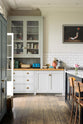 A closer look at a tall-ceilinged classic English kitchen centres around a wide painter’s black island with an aged brass Ionian tap, marble worktop, and glazed base cabinets. A cosy breakfast nook with vintage stools sits opposite an off-white cooker run and matching wall cabinets. The space feels artfully lived-in—funky paintings, and kitchen essentials tell the story of a creative, full-of-life family.