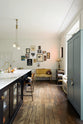 A tall-ceilinged classic English kitchen centres around a painter’s black island with a marble worktop, aged brass Ionian tap, and a full-height pantry cabinet. A cosy breakfast nook includes vintage stools, a gold-framed mustard sofa, and a white one by the window, draped in lace. Funky art, hanging sculptures, a vintage book trolley, and a delicate glass gaselier light give the space a creative, lived-in feel.