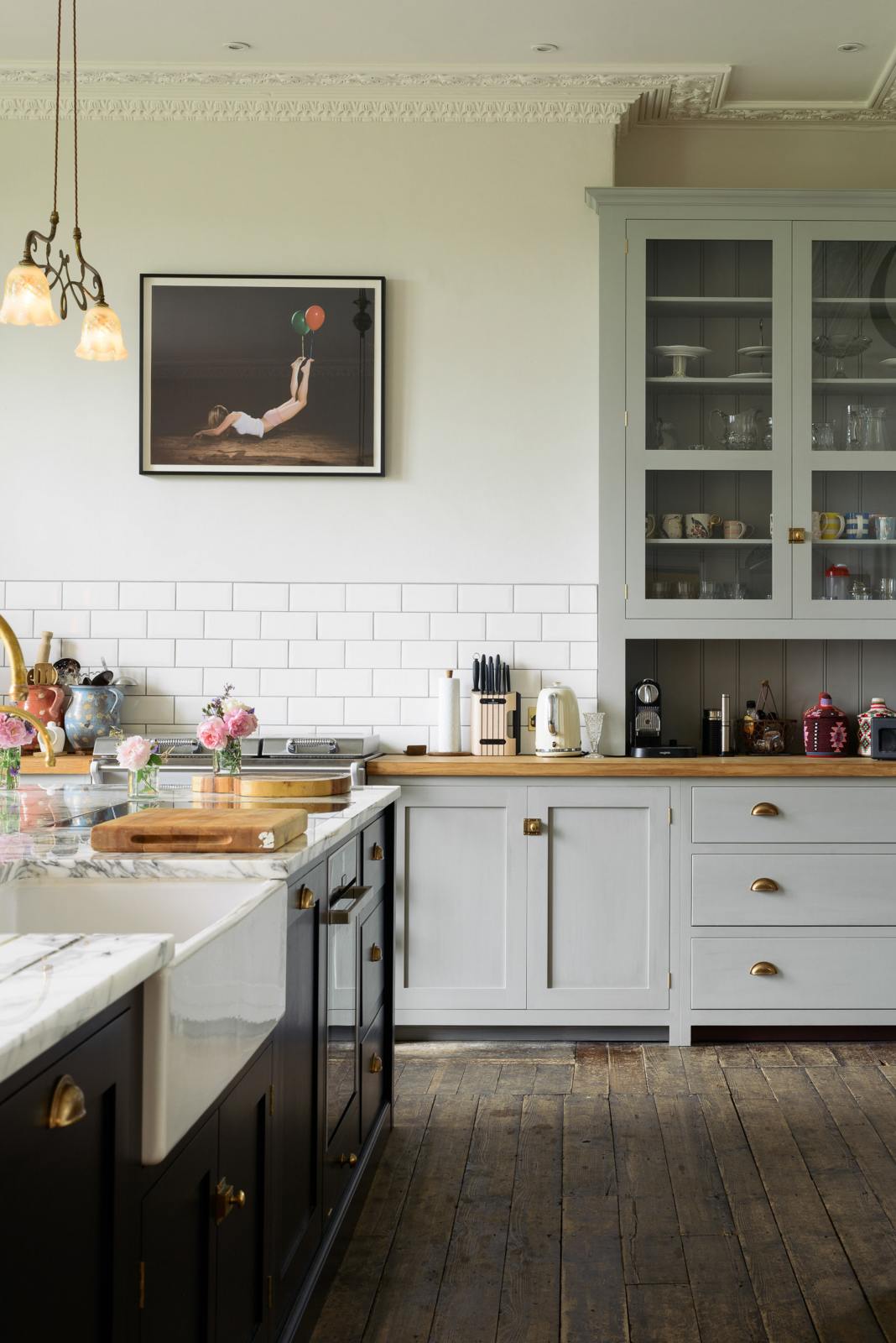 A tall-ceilinged, classic English kitchen features a wide painter’s black island with an aged brass Ionian tap, marble worktop, a cosy breakfast nook with vintage stools, an off-white cooker run, and matching wall cabinets. The space feels artfully lived-in — funky paintings, a delicate glass gaselier light, and kitchen essentials tell the story of a creative, lively family.