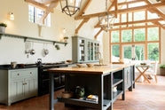 This oak-framed barn kitchen features a long cooker run with a black granite worktop, a bank of glazed larders, a shelf, and a kitchen island complete with a single Belfast sink and an aged brass Ionian tap atop a solid oak worktop. Adorned with framed pendant lights, artisan ceramics, books, and silver kitchenware, this kitchen creates a warm and inviting space.