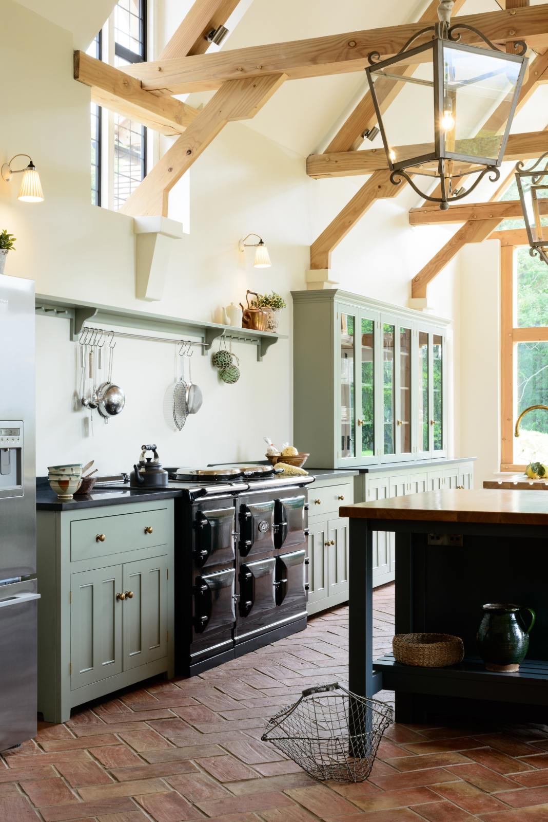 An oblique view of an oak-framed barn kitchen features a long cooker run with a black granite worktop, a bank of glazed larders, a shelf, and a kitchen island complete with a single Belfast sink and an aged brass Ionian tap atop a solid oak worktop. Adorned with framed pendant lights, artisan ceramics, books, and silver kitchenware, this kitchen creates a warm and inviting space.