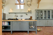 This oak-framed barn kitchen features a long cooker run with a black granite worktop, a bank of glazed larders, a shelf, and a kitchen island complete with a single Belfast sink and an aged brass Ionian tap atop a solid oak worktop. Adorned with framed pendant lights, artisan ceramics, books, wire baskets, and silver kitchenware, this kitchen creates a warm and inviting space.