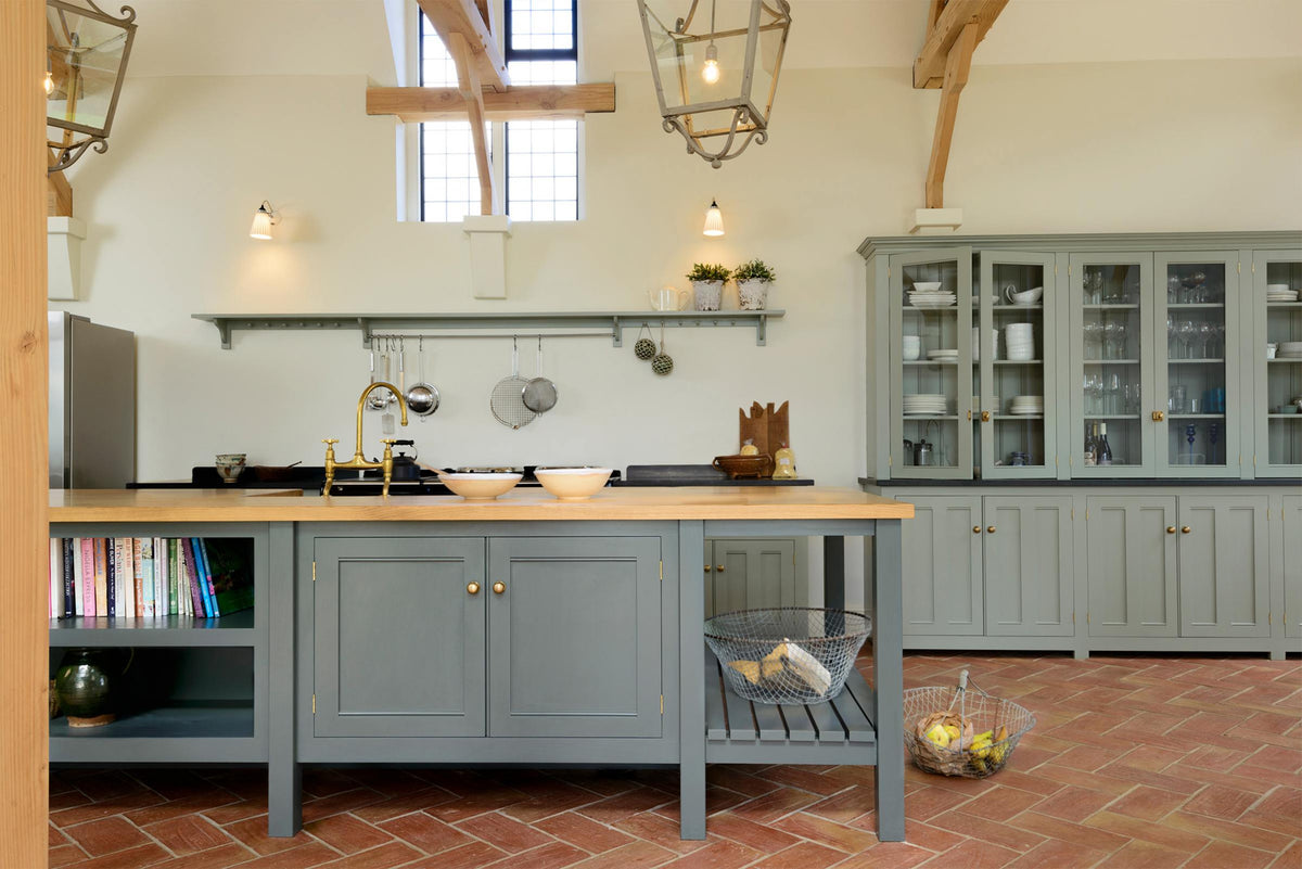 This oak-framed barn kitchen features a long cooker run with a black granite worktop, a bank of glazed larders, a shelf, and a kitchen island complete with a single Belfast sink and an aged brass Ionian tap atop a solid oak worktop. Adorned with framed pendant lights, artisan ceramics, books, wire baskets, and silver kitchenware, this kitchen creates a warm and inviting space.