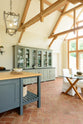 An oblique view of an oak-framed barn kitchen features a bank of glazed larders, a kitchen island complete with a single Belfast sink, and an aged brass Ionian tap atop a solid oak worktop. Adorned with framed pendant lights, a glass dining table, pears and pumpkins, this kitchen creates a warm and inviting space.
