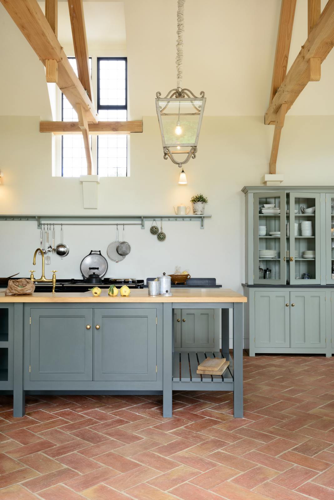 This oak-framed barn kitchen features a long cooker run with a black granite worktop, a shelf, and a kitchen island complete with a single Belfast sink and an aged brass Ionian tap atop a solid oak worktop. Adorned with framed pendant lights, artisan ceramics, books, pears, and silver kitchenware, this kitchen creates a warm and inviting space.