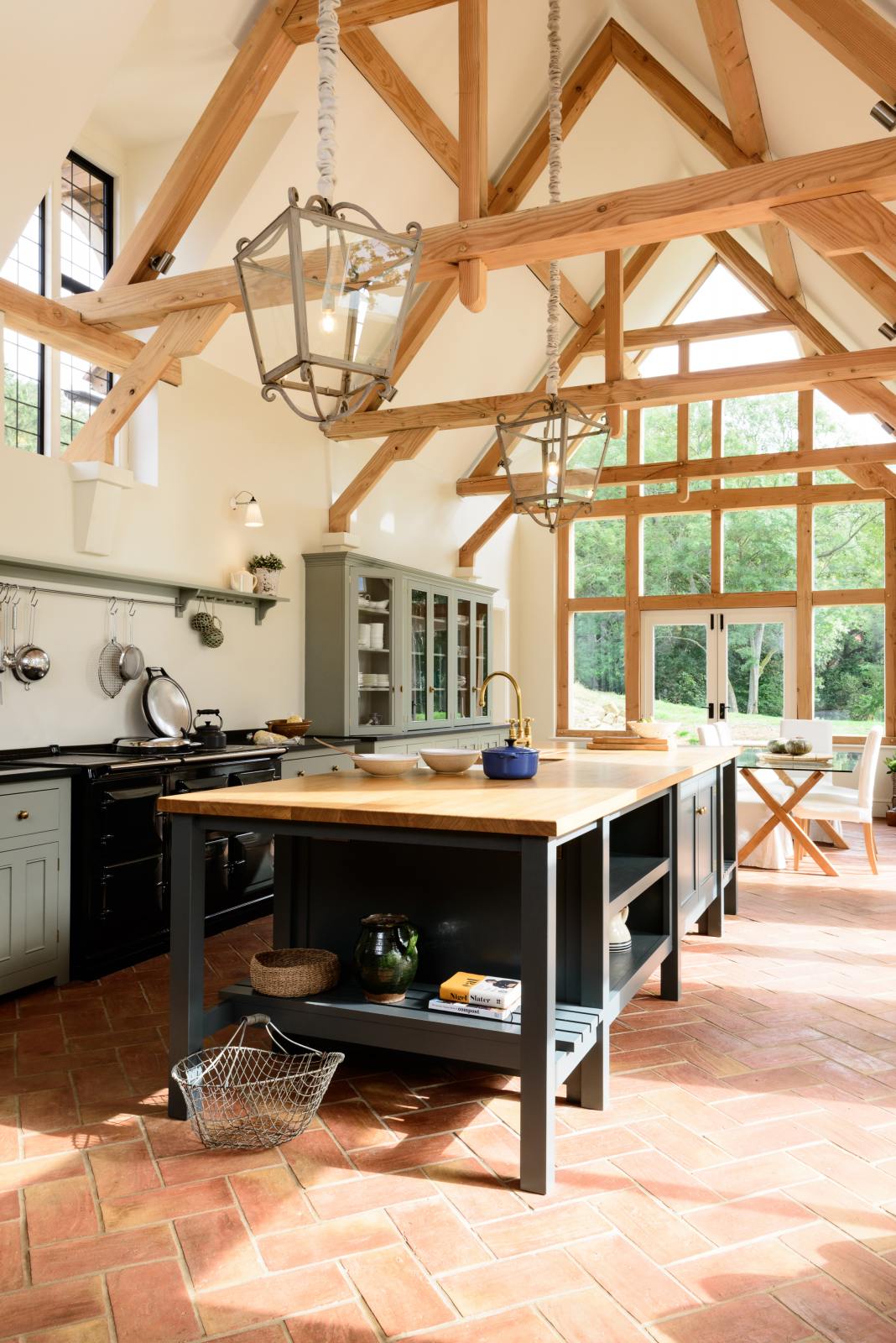 This oak-framed barn kitchen features a long cooker run with a black granite worktop, a shelf, and a kitchen island complete with a single Belfast sink and an aged brass Ionian tap atop a solid oak worktop. Adorned with framed pendant lights, artisan ceramics, books, pears, and silver kitchenware, this kitchen creates a warm and inviting space.
