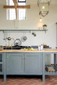 This oak-framed barn kitchen features a long cooker run with a black granite worktop, a shelf, and a kitchen island complete with a single Belfast sink and an aged brass Ionian tap atop a solid oak worktop. Adorned with framed pendant lights, artisan ceramics, books, pears, and silver kitchenware, this kitchen creates a warm and inviting space.