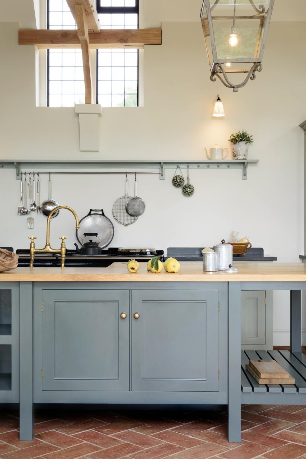 This oak-framed barn kitchen features a long cooker run with a black granite worktop, a shelf, and a kitchen island complete with a single Belfast sink and an aged brass Ionian tap atop a solid oak worktop. Adorned with framed pendant lights, artisan ceramics, books, pears, and silver kitchenware, this kitchen creates a warm and inviting space.