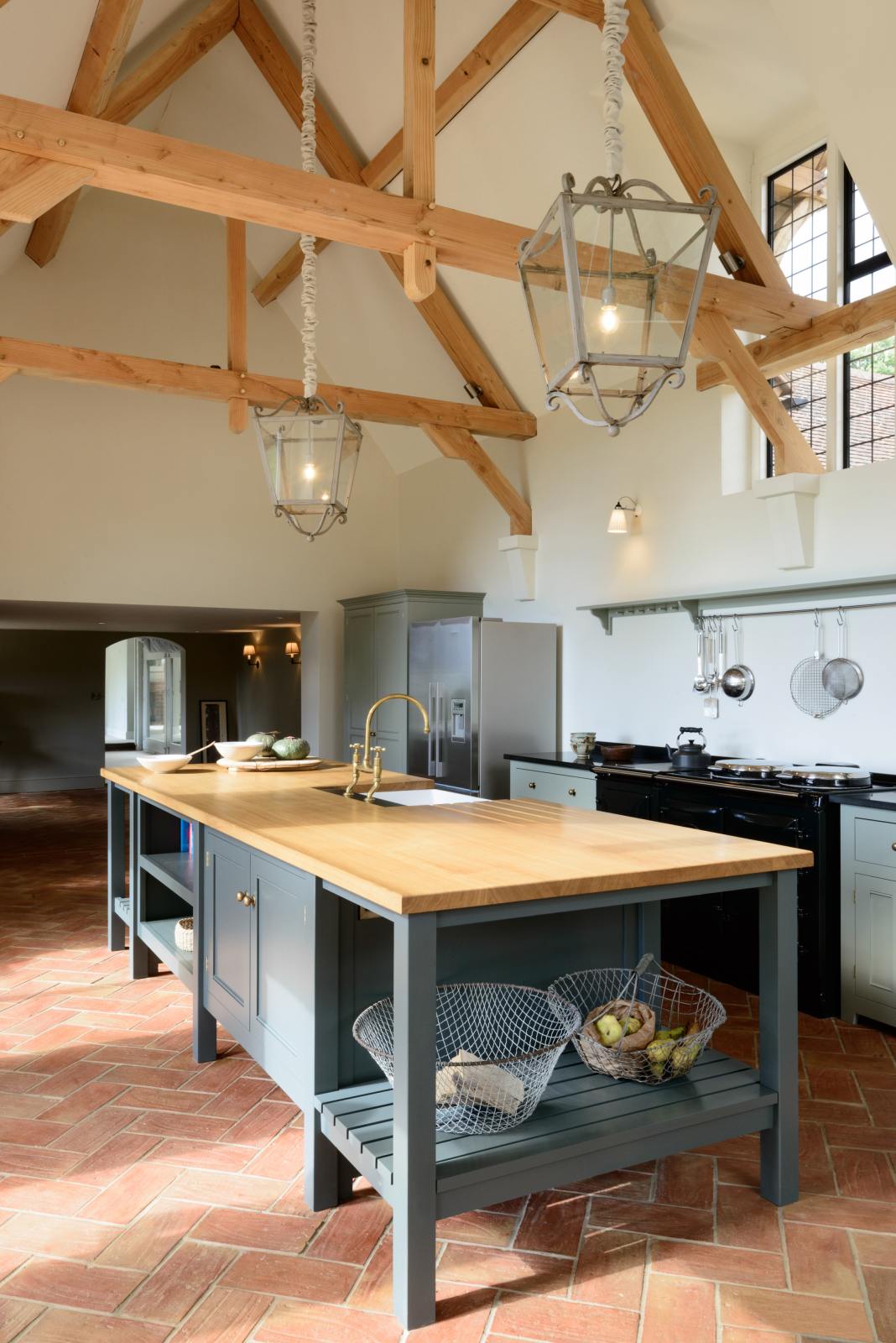 An oblique view of an oak-framed barn kitchen features a long cooker run with a black granite worktop, a shelf, and a kitchen island complete with a single Belfast sink and an aged brass Ionian tap atop a solid oak worktop. Adorned with framed pendant lights, wired baskets, fruits, and silver kitchenware, this kitchen creates a warm and inviting space.