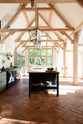 An oblique view of an oak-framed barn kitchen features a long cooker run with a black granite worktop, a bank of glazed larders, a shelf, and a kitchen island complete with a single Belfast sink and an aged brass Ionian tap atop a solid oak worktop. Adorned with framed pendant lights, artisan ceramics, books, and pumpkins, this kitchen creates a warm and inviting space.