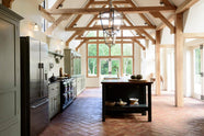 An oak-framed barn kitchen features a long cooker run with a black granite worktop, a bank of glazed larders, a shelf, and a kitchen island complete with a single Belfast sink and an aged brass Ionian tap atop a solid oak worktop. Adorned with framed pendant lights, artisan ceramics, books, and pumpkins, this kitchen creates a warm and inviting space.