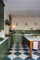 A spacious and elegant Wilkes green classic English kitchen features a long L-shaped cooker and sink run with a Silestone worktop, glazed wall cupboards, shelves, and an Heirloom dairy table. Adorned with classic marble chequered-board flooring, Heirloom task lights, vintage ceramics, copper pots, and luscious green vegetables, it creates a classic and stylish kitchen.