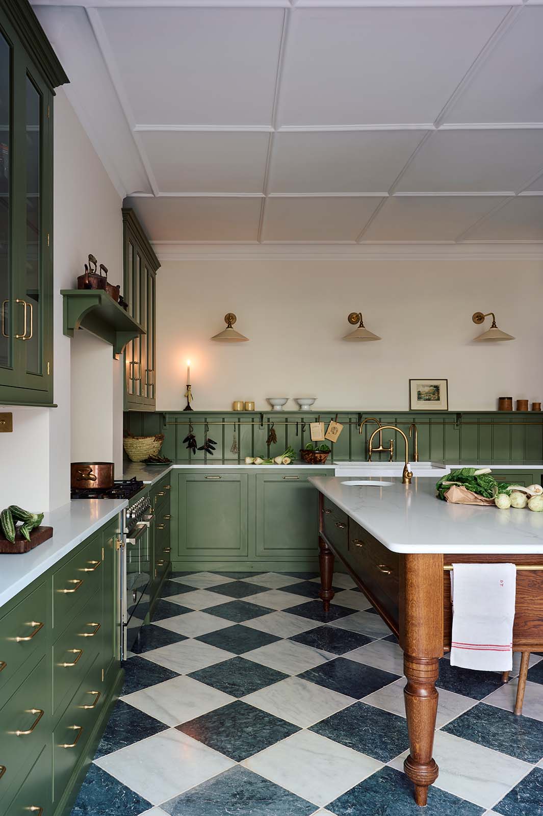 A spacious and elegant Wilkes green classic English kitchen features a long L-shaped cooker and sink run with a Silestone worktop, glazed wall cupboards, shelves, and an Heirloom dairy table. Adorned with classic marble chequered-board flooring, Heirloom task lights, vintage ceramics, copper pots, and luscious green vegetables, it creates a classic and stylish kitchen.