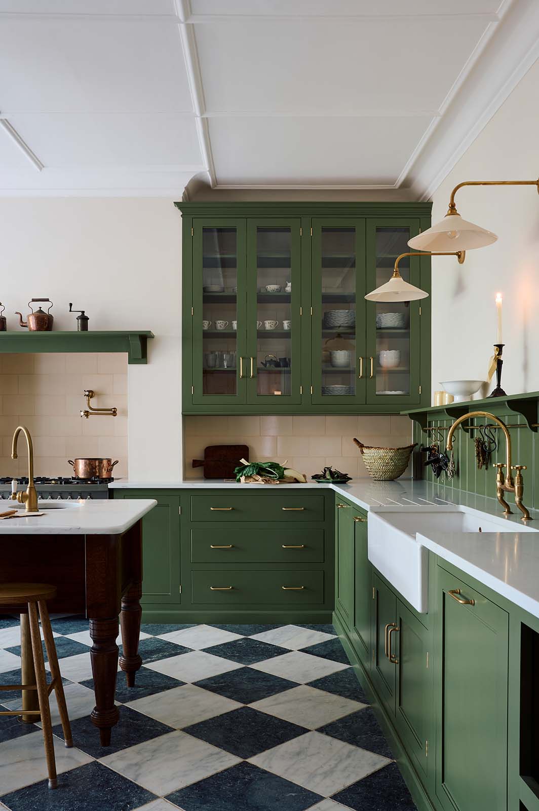 A spacious and elegant Wilkes green classic English kitchen features a long L-shaped cooker and sink run with a Silestone worktop, glazed wall cupboards, shelves, and an Heirloom dairy table. Adorned with classic marble chequered-board flooring, Heirloom task lights,  cream subway splashback tiles, copper pots, and luscious green vegetables, it creates a classic and stylish kitchen.
