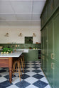 A spacious and elegant Wilkes green classic English kitchen features a long L-shaped cooker and sink run with a Silestone worktop, a bank of full-height pantry cupboards, a long shelf, and an Heirloom dairy table. Adorned with classic marble chequered-board flooring, Heirloom task lights, vintage ceramics, Helen stools, and luscious green vegetables, it creates a classic and stylish kitchen.