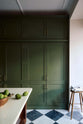 A spacious and elegant Wilkes green classic English kitchen features a bank of full-height pantry cupboards, and an Heirloom dairy table. Adorned with classic marble chequered-board flooring, a bowl of limes, a Helen stool, and tea towels, it creates a classic and stylish kitchen.