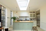 This bright and spacious classic English kitchen features a long white cooker run with glazed wall cupboards and a pale-blue kitchen island that includes a single Belfast sink, an aged brass Ionian tap, and a solid oak worktop. Adorned with metal embellishments like industrial pendant lights, a silver refrigerator, a jade-green vase filled with a floral arrangement, and vintage wooden furniture, it creates a wonderful mix of simplicity and charismatic personality.