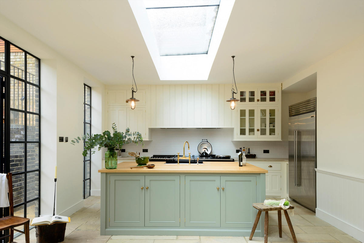 This bright and spacious classic English kitchen features a long white cooker run with glazed wall cupboards and a pale-blue kitchen island that includes a single Belfast sink, an aged brass Ionian tap, and a solid oak worktop. Adorned with metal embellishments like industrial pendant lights, a silver refrigerator, a jade-green vase filled with a floral arrangement, and vintage wooden furniture, it creates a wonderful mix of simplicity and charismatic personality.