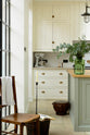 A quite corner of a bright and spacious classic English kitchen features a long white cooker run with wall cupboards and a pale-blue kitchen island with a solid oak worktop. Adorned with metal embellishments like industrial pendant lights, a silver mixer, a jade-green vase filled with a floral arrangement, and vintage wooden furniture, it creates a wonderful mix of simplicity and charismatic personality.