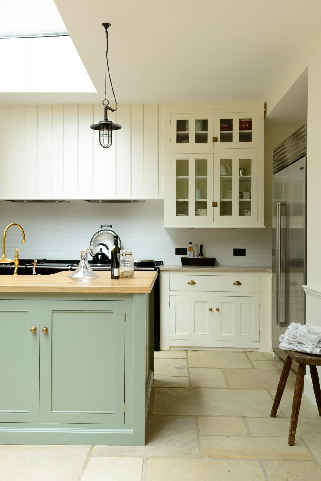 A quiet corner of a bright and spacious classic English kitchen features a long white cooker run with glazed wall cupboards and a pale-blue kitchen island that includes a single Belfast sink, an aged brass Ionian tap, and a solid oak worktop. Adorned with metal embellishments like industrial pendant lights, a silver refrigerator, a jade-green vase filled with a floral arrangement, and vintage wooden furniture, it creates a wonderful mix of simplicity and charismatic personality.